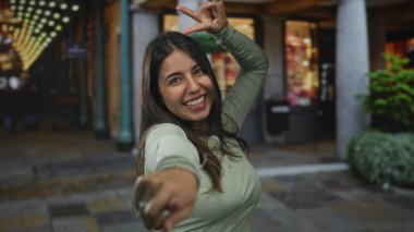 Akşam saatlerinde aydınlık bir şehir bölgesinde neşeli bir şekilde poz veren genç İspanyol kadın hareketli sokak hayatı ve dinamik duruş sergiliyor..