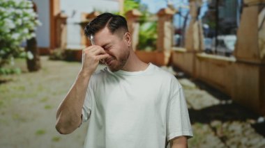 Parkta sakallı genç adam yorgun görünüyor. Burun köprüsü çimdikliyor, etrafı güneşli açık hava manzarasıyla çevrili. Beyaz bir gömlek giyiyor..