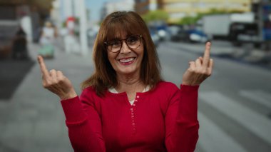 Gözlüklü ve kırmızı tişörtlü yaşlı kadın hareketli şehir caddesinde cesur bir jest yapıyor. Kendine güvenen tavır sergiliyor. Bulanık arabaları ve yayaları olan farklı şehirlerde yaşıyor.