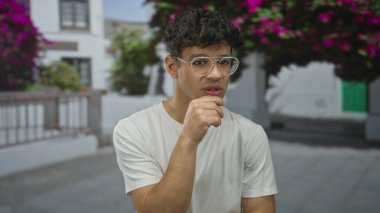 İspanyol gözlüklü adam eski kasaba caddesinde dururken çeneye dokunuyor; düşünceli bir düşünce anlayışı var..