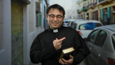 Şehir sokaklarında elinde kitap tutan, haç takan ve şehir ortamında neşeli bir ifade sergileyen bir adam.