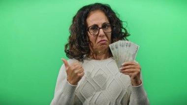 Kafası karışmış bir ifadeyle Dirham banknotları tutan ve sağlam yeşil bir zemine karşı baş parmak kaldıran bir kadın belirsizlik ve mali karar alma duyusunu iletiyor..