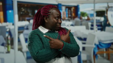 Yeşil kazaklı bir kadın açık hava restoranındaki terasta esprili bir şekilde el kol hareketi yapıyor, etkileyici vücut dili ve yüz ifadeleri sergiliyor..