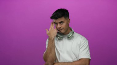 Kulaklıklı genç İspanyol bir adam pembe arka planda silah hareketi yapıyor, beyaz bir gömlek giyiyor ve etkileyici bir yüz ifadesi sergiliyor..