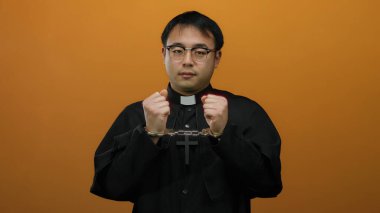 Rahip kıyafeti ve kelepçe takan Asyalı bir adam turuncu arka planda duruyor..