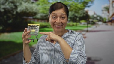 Orta yaşlı İspanyol bir kadın, elinde el arabası ve parmaklarıyla caddeyi işaret ediyor. Mutluluk alışverişi..