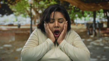 Açık hava parkında, ağaç gölgesinin altında şaşkınlık ve duygu gösteren, şok olmuş yüz ifadesi olan genç ve iri bir kadın.