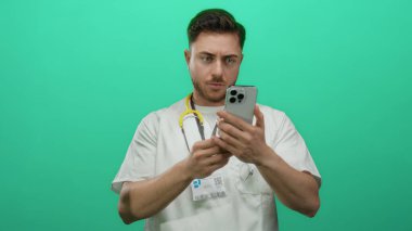 Beyaz önlüklü genç sakallı doktor, izole edilmiş yeşil arka plana dikkatle bakıyor, steteskop ve kimlik kartı takıyor, modern sağlık hizmetleri profesyonelini somutlaştırıyor..