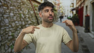 Taş duvar ve bulanık yayalar ile dar bir sokakta göğsü gösteren genç adam parmağı; belirsizlik şüphesi kafa karışıklığı.