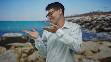 Beyaz gömlekli adam gülümsüyor ve stüdyoda kayalık kıyılarda avuçlar açık, mavi deniz ise iyimser..