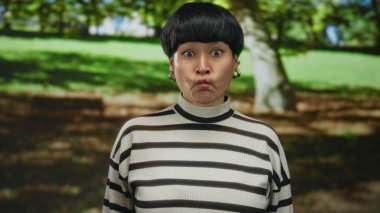 Genç bir kadın açık hava parkında, yemyeşil ağaçların arasında komik bir yüz ifadesi sergiliyor, neşeli ifadeler sergiliyor ve doğal bir çevrede enerjik bir enerji sergiliyor..