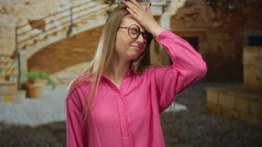 Güneşli bir şehir bölgesinde pembe gömlekli, yuvarlak gözlükle poz veren kadın, bulanık tarihi bir arkaplan karşısında çeşitli ifadeler sergiliyor, unutkanlık ya da farkındalık öneriyor..