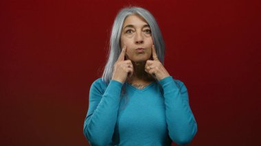 Kıdemli kadın mavi kazaklı, kırmızı duvarın arkasında neşeli karakterini vurgulayarak neşeli bir yüz ifadesi takınıyor..