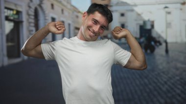 Güler yüzlü İspanyol adam gündüz vakti şehir meydanındaki güneş ışığıyla aydınlanan kaldırım taşı caddesinde yanak jesti yapıyor..