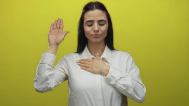 Beyaz tişörtlü, eli yüreğinde genç İspanyol bir kadın canlı sarı arka plana karşı poz veriyor. Samimiyet ve bağlılık gösteriyor.