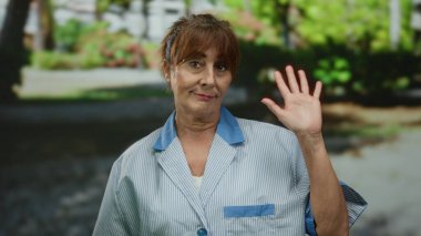 Kıdemli İspanyol kadın parkta jest yaparken gülümsüyor, güneşli bir günde dışarıda üniforma giyiyor..