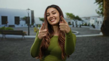 Beyaz binalar ve arka planda yeşil alanlar olan bir sokak ortamında gülümseyen ve şakacı bir şekilde kamerayı işaret eden genç İspanyol kadın..