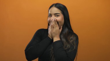 Elleri ağzının üzerinde gülen bir kadın canlı turuncu bir duvarın karşısında neşeli, neşeli bir atmosferi olan, neşeli ve izole bir ortamda neşe ve şaşkınlığı ifade ediyor..