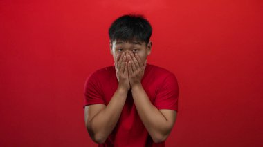 Şaşırmış ifadesi olan genç bir adam, izole bir arka planda parlak kırmızı bir duvara karşı elleri ile ağzını kapatır.