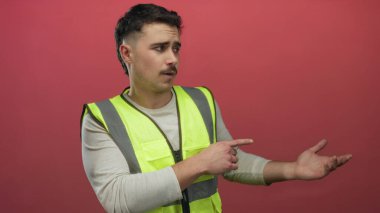 Yansıtıcı yelek giymiş genç İspanyol bir adam, izole edilmiş kırmızı arka plan duvarına işaret ediyor, odaklanmış bir ifadeyle yönü gösteriyor..