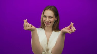 Genç beyaz kadın, mor bir stüdyo ortamında parmaklarını sıkarken neşeli bir şekilde gülümsüyor..