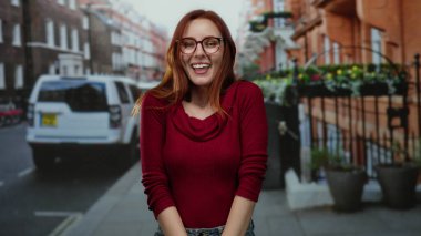 Kırmızı kazaklı kadın arka planda arabalar ve binalarla şehir caddesinde duruyor, gülümsüyor ve kameraya bakıyor, güneşli bir gün, günlük kıyafetler.