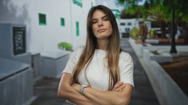 Şehir caddesinde kendine güvenen genç bir kadın, kollarını kavuşturmuş, gündelik kıyafetler giymiş, modern şehir mimarisi ve açık hava ortamında yeşil ağaçlarla çevrili..