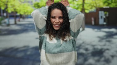 Genç bir kadın, güneşli bir günde arka planda ağaçlarla dışarıda dikilirken elleri kafasında tavşan kulağı hareketi yapıyor..