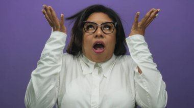 Gözlüklü kadın, mor arka planda ellerini kaldırmış, şok halinde. Beyaz bir gömlek giyiyor. Duygularını ve şaşkınlığını canlı bir arkaplanda aktarıyor..