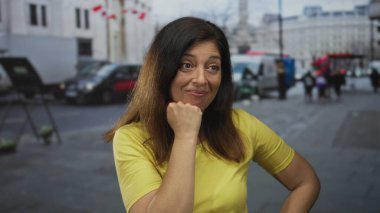 Orta yaşlı, sarı tişörtlü, elleri çenesinde, trafiğin yoğun olduğu bir ortamda şehir sokağında düşünceli poz veren İspanyol bir kadın..
