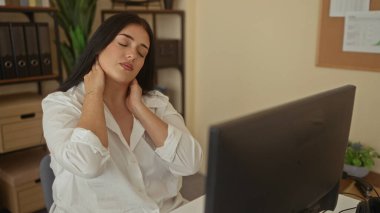 Ofisteki kadın, ofis ortamındaki bir bilgisayarın başında oturmuş, organize dosyalar ve ofis malzemeleriyle çevrili bir şekilde çalışırken boynunu sıvazlıyor..