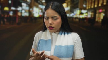 Dışarıda parmaklarını sayan bir kadın var. Yoğun bir caddede arka planı bulanık. Beyaz tişört ve mavi eşarp giyiyor..