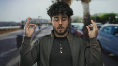 Sakallı genç adam güneşli gökyüzünün altında meditasyon yapıyor. Bulanık araba ve ağaçların arasında huzurlu bir manzara yaratarak huzurlu bir yüz ifadesi sergiliyor.