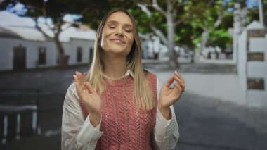 Güneşli bir günde açık havada, sakin bir sokak arkaplanıyla gülümseyen, pembe bir kazak ve beyaz gömlek giyen bir kadın, şehir ortamının tadını çıkarırken neşeli ve rahat görünüyor..
