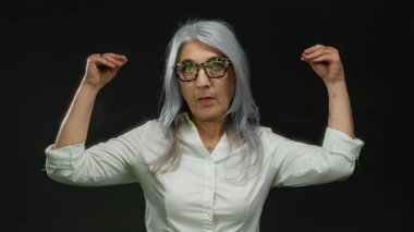 Tutkulu jestler, gri saçlar ve gözlüklerle konuşan, siyah bir arkaplanda izole edilmiş, duygularını ve bağlılığını ifade eden bir kadın..