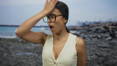 Kayalık bir sahilde gözlüklü bir kadın, deniz kenarında şaşkınlık ve farkındalık ifadesi takınıyor..