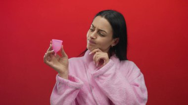 Pembe bornozlu bir kadın canlı kırmızı arka planda, canlı kırmızı bir arka planda regl bardağını inceliyor. Merakını, dalgınlığını ve kişisel bakımını canlı ve izole bir ortamda sergiliyor..