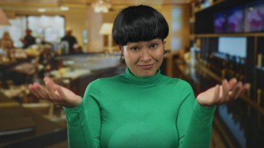 Restoran ortamında kapalı alanda el kol hareketi yapan yeşil gömlekli bir kadın, parmakları tırnak içinde şüphecilik taslayan, kafeterya atmosferinin yumuşak bulanık arka planına sahip bir kadın..