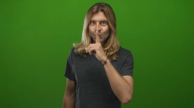 Koyu gri gömlekli sarışın adam yeşil stüdyoda sessizlik için parmaklarını dudaklarına işaret ediyor..