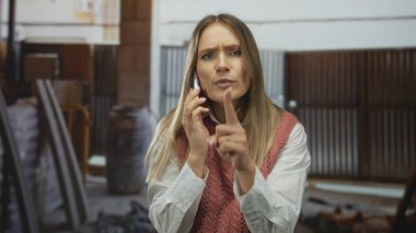 Sarı saçlı kadın akıllı telefondan konuşuyor ve kapalı bir inşaat sahasında sessizlik için işaret veriyor, mahremiyeti vurguluyor..