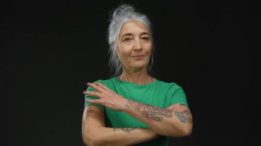 Gri saçlı ve dövmeli yaşlı bir kadın siyah izole bir arka plana karşı kollarını kavuşturarak duruyor..