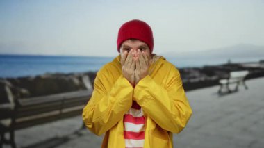 Kırmızı bereli ve sarı yağmurluklu genç adam arka planda okyanus manzaralı deniz kenarında gezinti yapmaktan zevk alıyor..