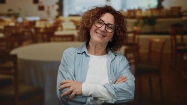 Konforlu bir restoran ortamında kollarını kavuşturup gülümseyen bir kadın, gündelik güven ve dostça davranışlar sergiliyor..