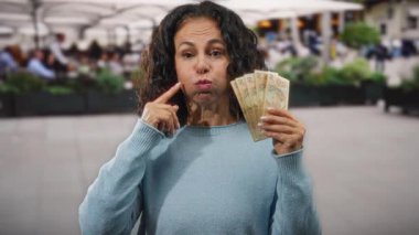 Kafe terasında orta yaşlı bir kadın elinde Brezilya banknotları tutarak şakacı yanaklar şişiriyor. Çevresindeki farklı insanlar ve şehir geçmişiyle neşeli bir sahne yaratıyor..