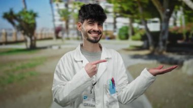 Parkta gülümseyen ve el kol hareketi yapan genç bir adam. Arka planda ağaçlar ve binalar olan beyaz bir ceket giyiyor. Açık havada dostane ve cana yakın bir tavır sergiliyor..