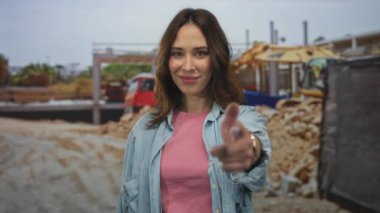 İspanyol kadın inşaat alanında gülümsüyor, günlük kıyafetler giyiyor, bulanık bina makineleri ve açık hava düzeneği ile canlı, genç bir atmosferi taşıyor..