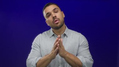 Latin asıllı genç adam dua jesti ile çıplak ellerini birbirine bastırıyor ve mavi stüdyo ortamında gözlerini kapıyor; sakin yansıma.