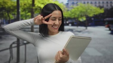 Şehir sokaklarında elinde bir defter olan genç bir İspanyol kadın. Etrafı ağaçların arasından süzülen şehir manzarası ve güneş ışığıyla çevrili bir barış hareketiyle gülümsüyor..