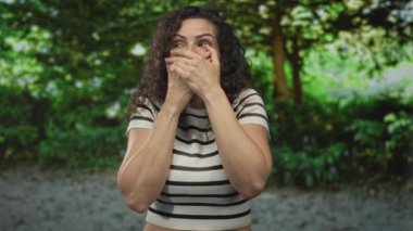 Kadının ağzı iki eliyle de gür yeşil ormanlarda görünür kıvırcık saçlarla, kaşları kalkık bir şekilde jest yapıyor..