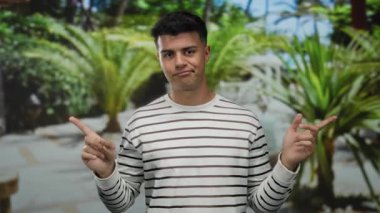 Genç adam parmaklarıyla bir açık hava parkı ortamında, yemyeşil ile çevrili, çizgili bir kazak giyiyor, oyuncu ve kendinden emin bir tavır sergiliyor..